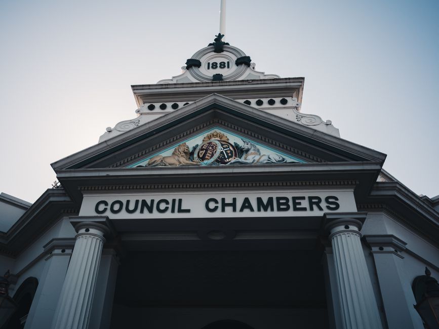 Photograph of the front façade of a council chambers building with classical architectural features, including white columns supporting a triangular pediment decorated with a colourful coat of arms an