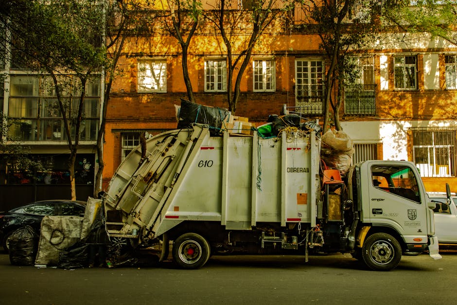 A person wearing orange work overalls standing inside a property, holding two large blue recycling bags filled with waste materials, one in each hand, during the home relocation or packing process. Th