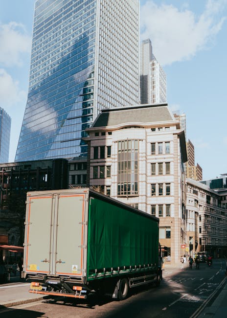 A light blue removal van is parked on a busy city street in front of historic multi-storey buildings with a mix of brick and ornate architectural details, including arched windows and decorative facad
