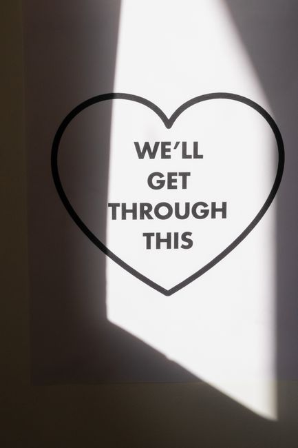 A white wall with a large black outline of a heart, inside which the words 'WE'LL GET THROUGH THIS' are printed in bold, black capital letters. Light from a nearby window casts a rectangular shadow ac