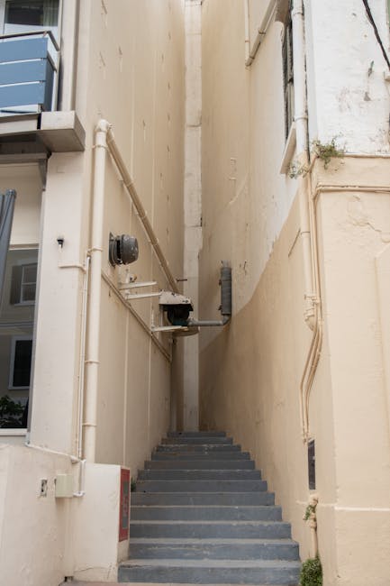 Exterior view of a building entrance with a short flight of concrete stairs leading up to a dark grey door, surrounded by grey stone walls and a black metal gate at the bottom of the stairs. The gate 